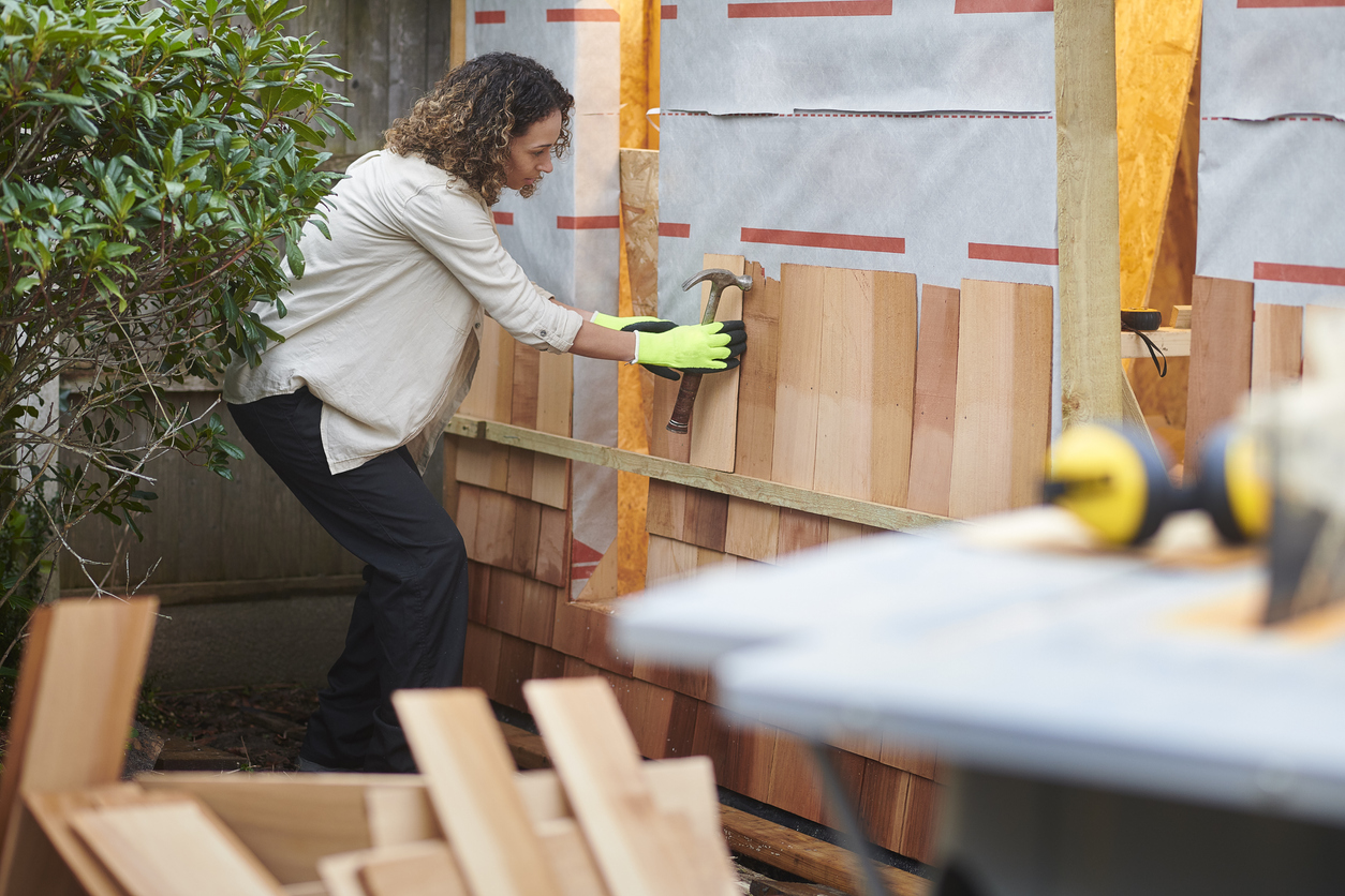 woman-works-on-a-wooden-shed-while-holding-a-hammer-and-wearing-gloves