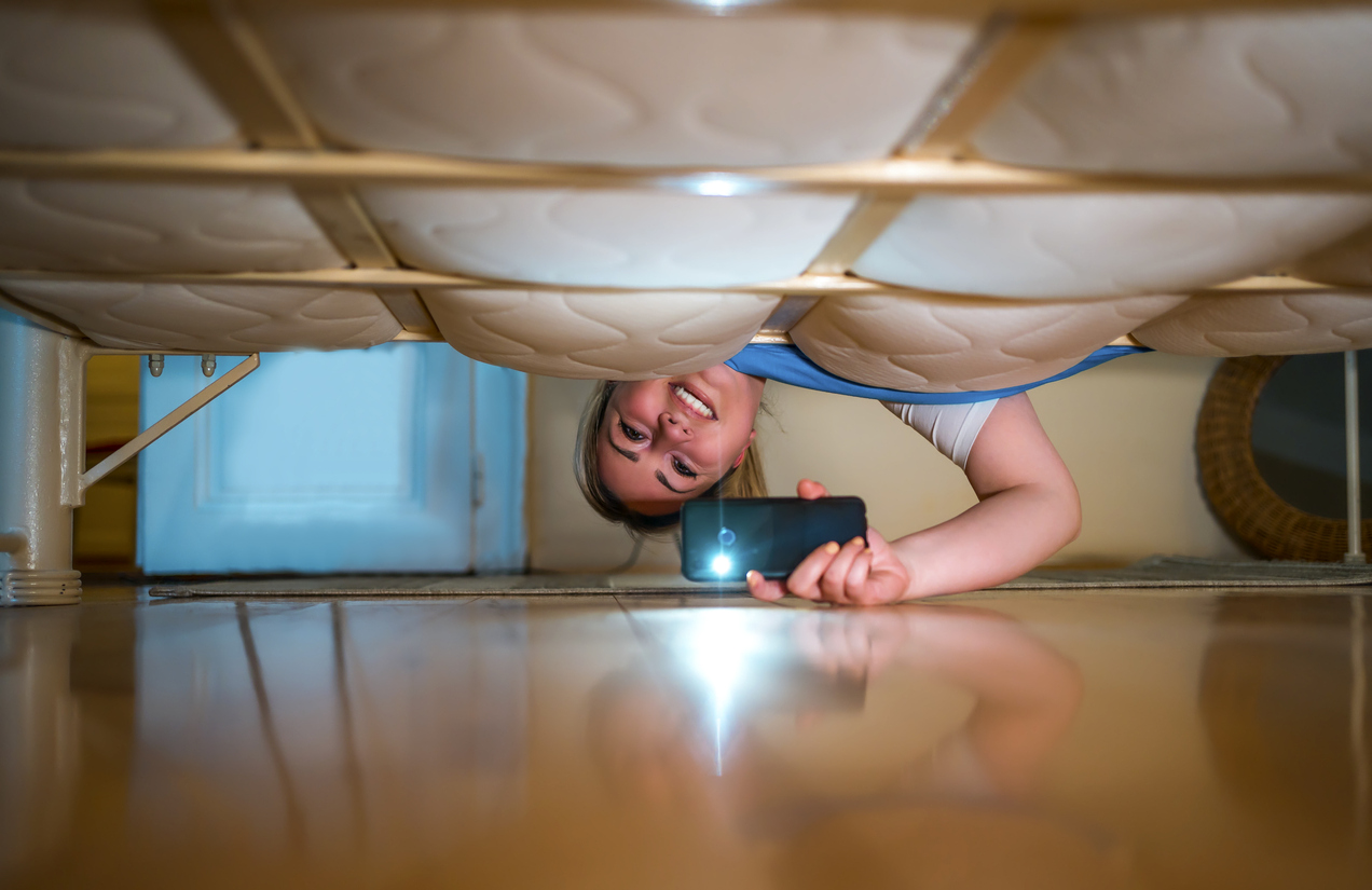 Young smiling woman looking under the bed, using flashlight in search