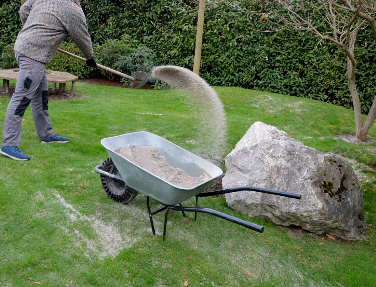 Person shoveling sand from a wheelbarrow onto a lawn.