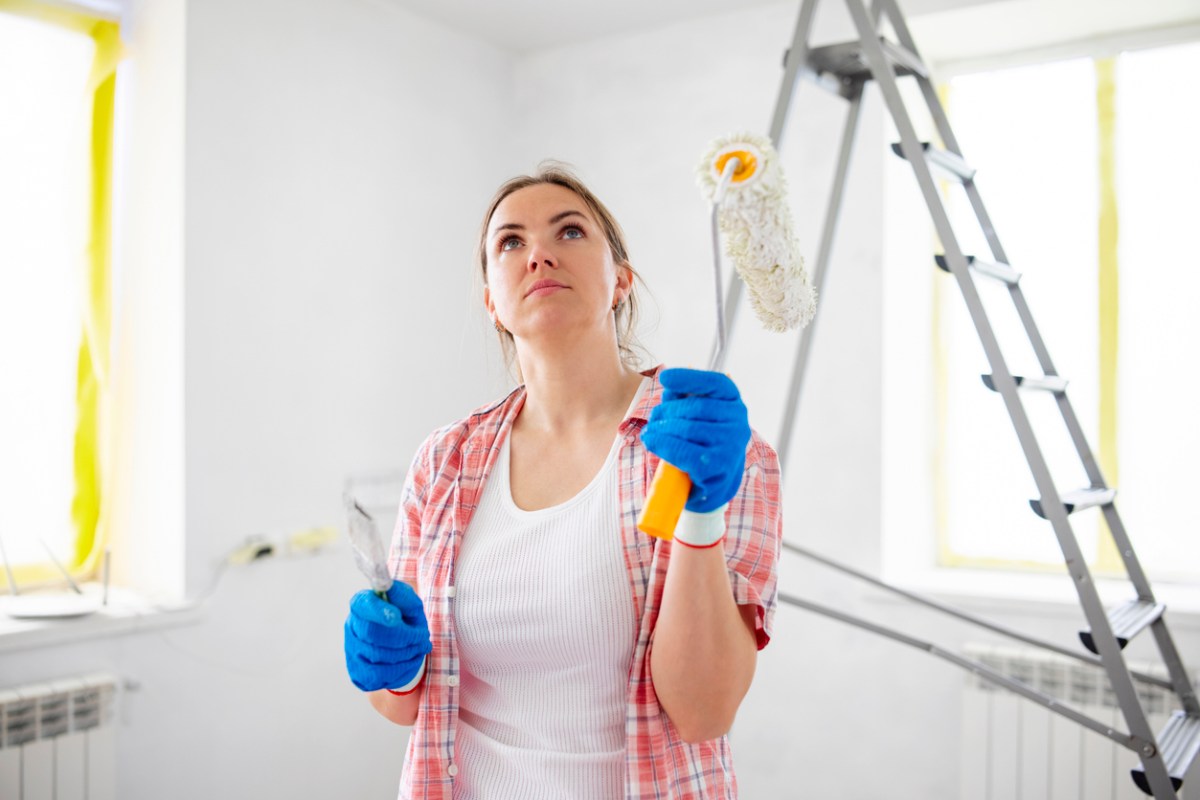 woman in room with ladder and paint supplies holding a paint roller and looking at the wall she is painting