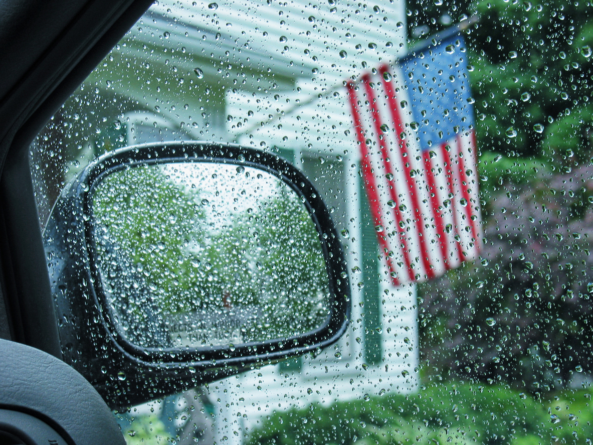 view from inside a car of an American flag hanging from a house with the windows and rear view mirror covered in rain drops