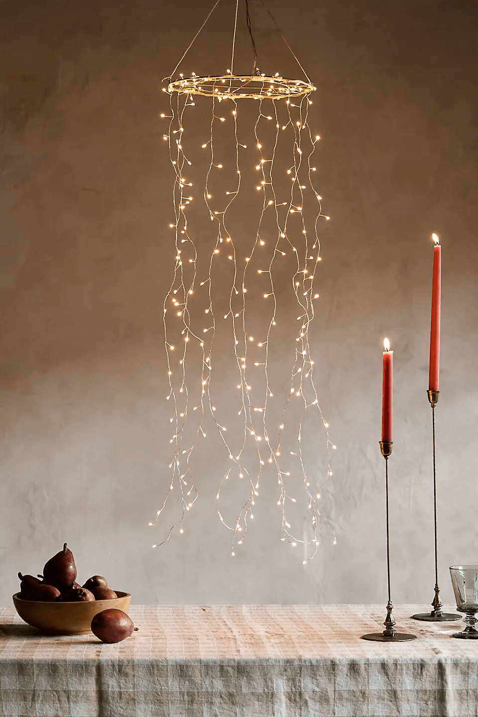 A-cascading-string-light-chandelier-hangs-over-a-table-with-fruit-and-candle-sticks.