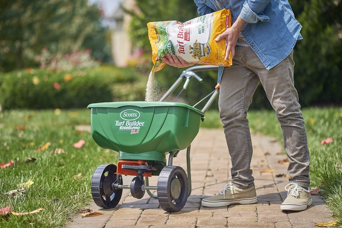 A person pouring a large bag of the best winter grass fertilizer into a push spreader that's sitting on a bricked walkway next to a lawn