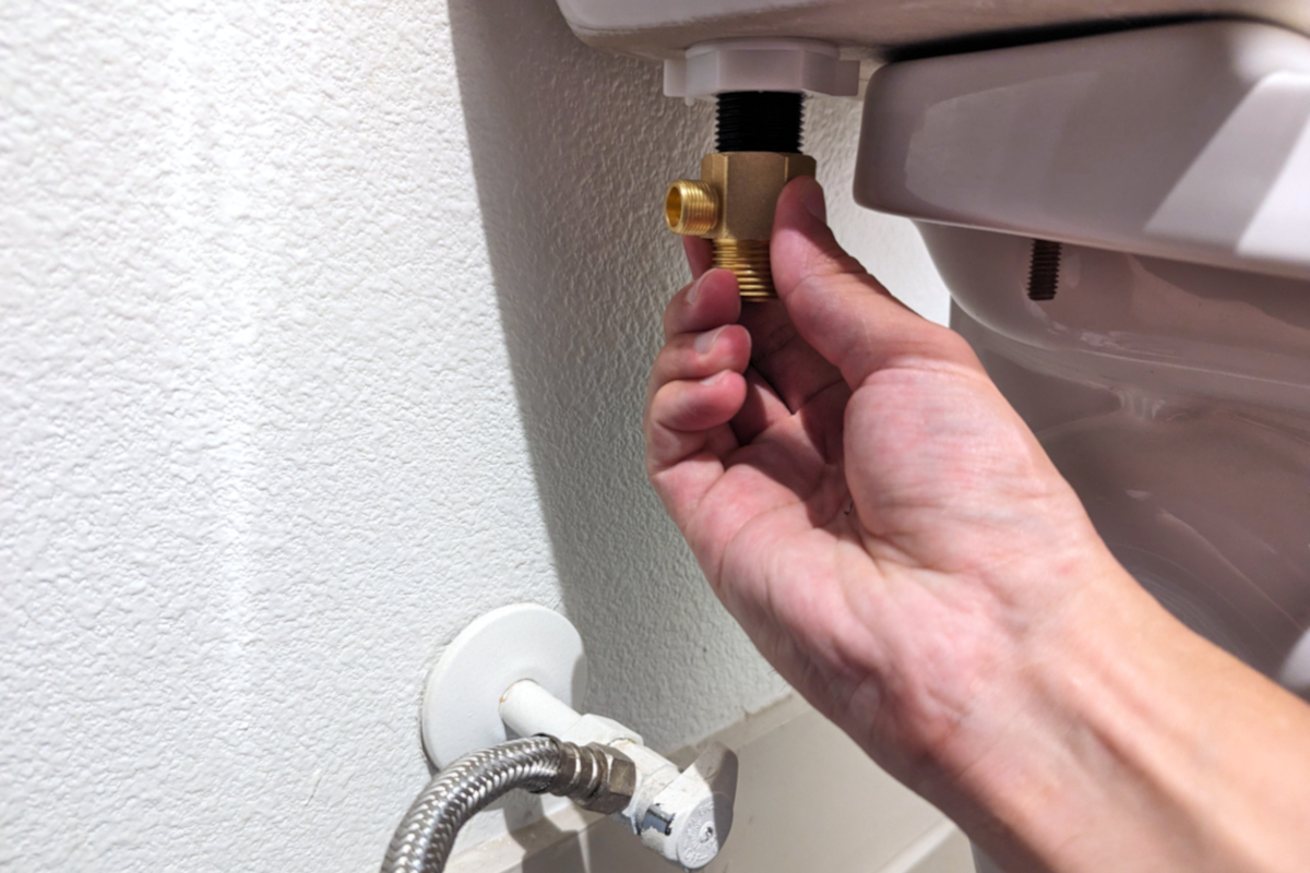 A person tightening a brass bidet T-adapter onto the base of their toilet tank by hand.