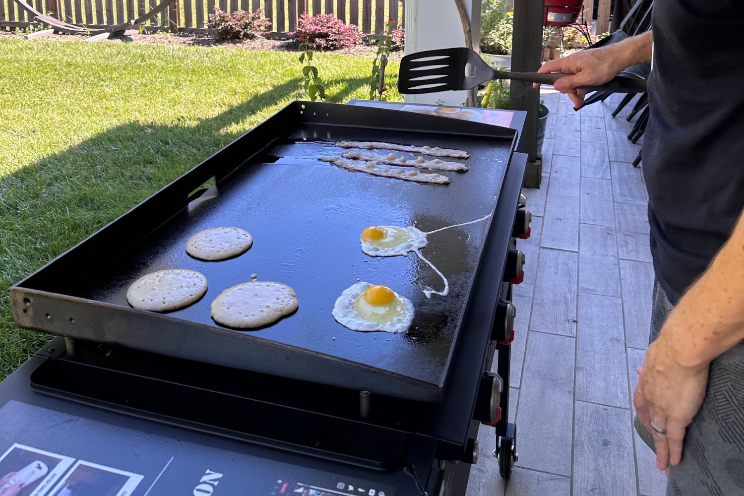 Person cooking pancakes, eggs, and bacon on 36-inch Blackstone griddle in backyard.