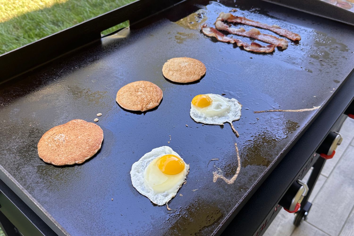 Three pancakes, two eggs, and three strips of bacon cooking on a griddle.