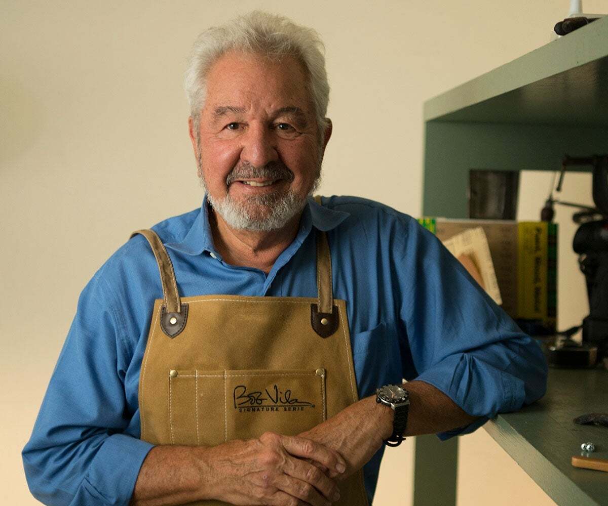 Bob Vila with grey hair and beard smiles while wearing overalls in a workshop.