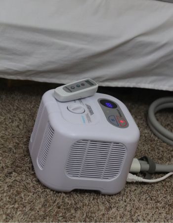 The Chilipad Sleep System and remote sitting on a carpeted floor next to a bed.