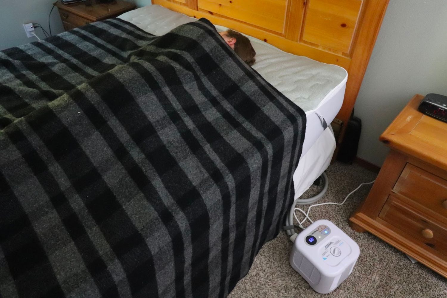 A person under a plaid blanket in a bed that has the Chilipad Sleep System installed on it.