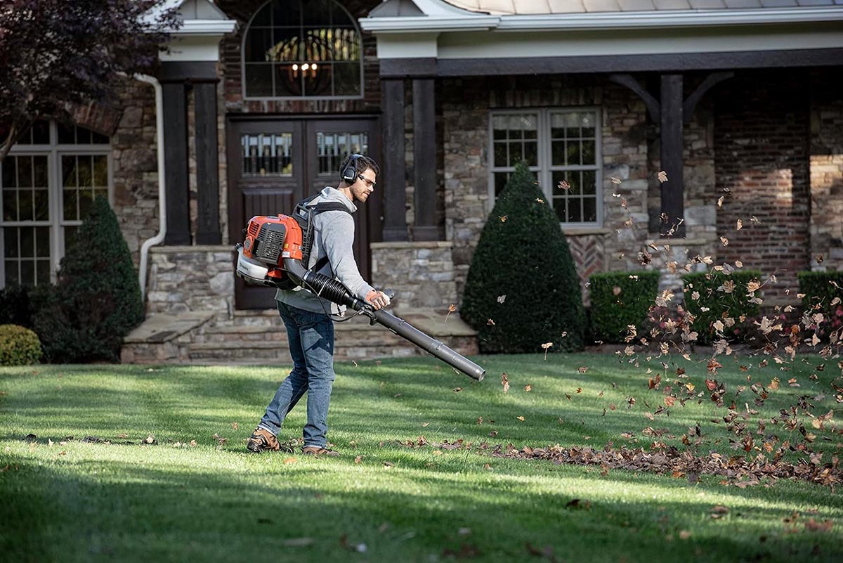A person using a Husqvarna backpack leaf blower to move leaves in a yard with a large house in the background.