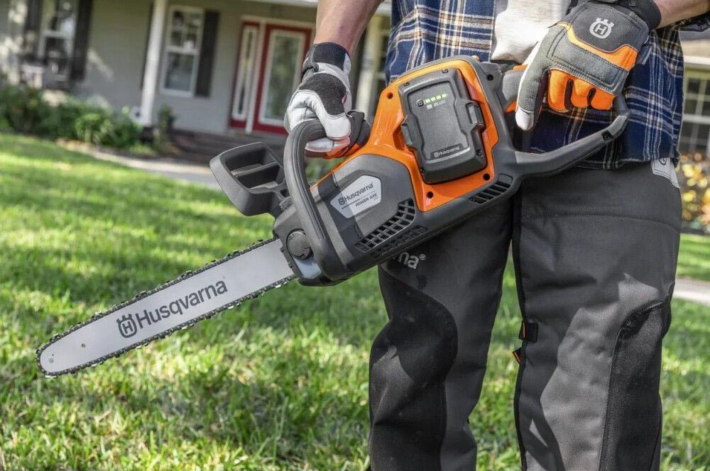 A person holding a Husqvarna chainsaw while standing in a yard in front of a house.