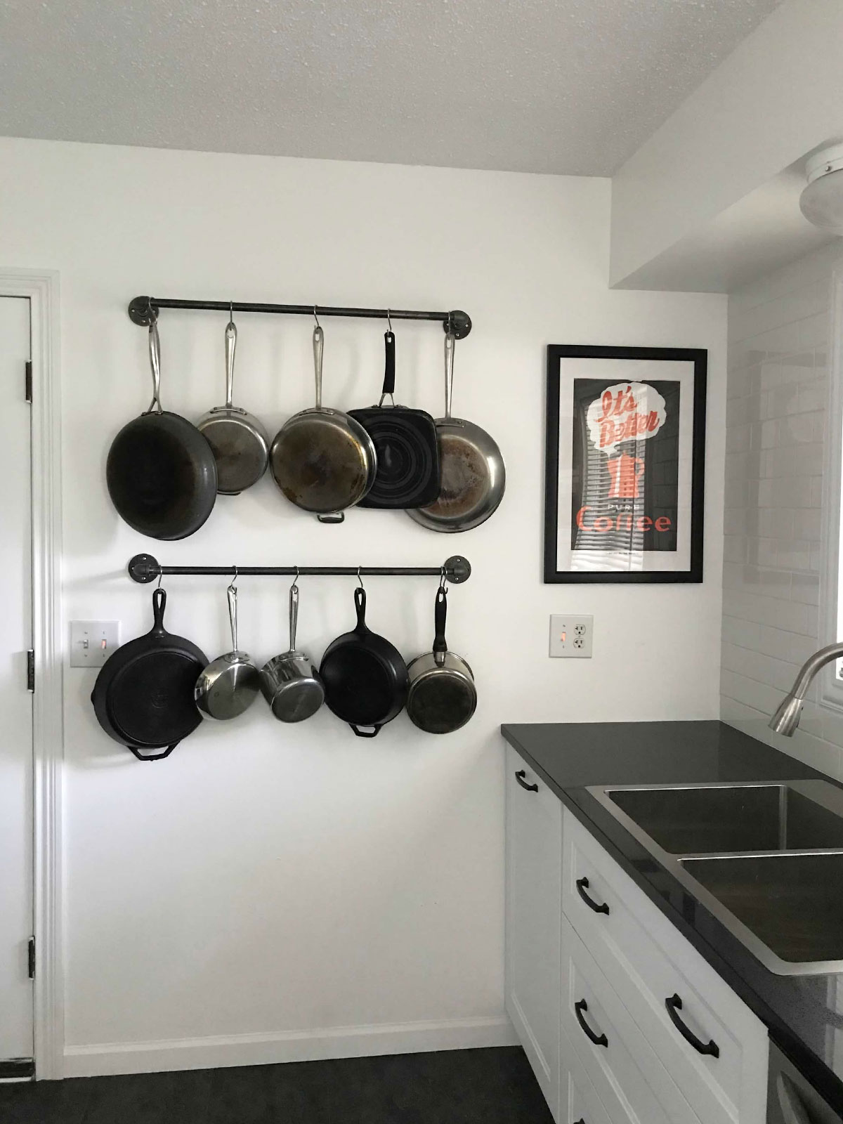 A pot rack against a white wall in a white and black kitchen displays a variety of pans and pots.