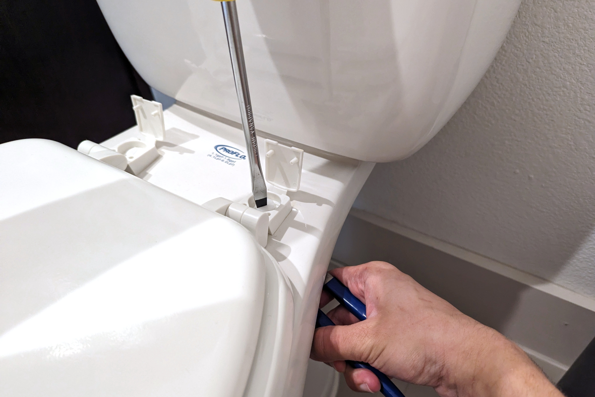 Person unscrewing a toilet seat bolts using a flathead screwdriver and channel lock pliers on a white two-piece ProFlo toilet