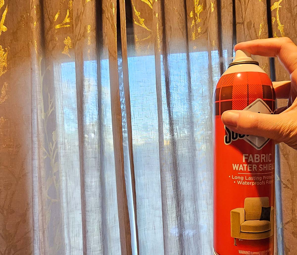 A person spraying Scotchgard fabric protector on hanging curtains.