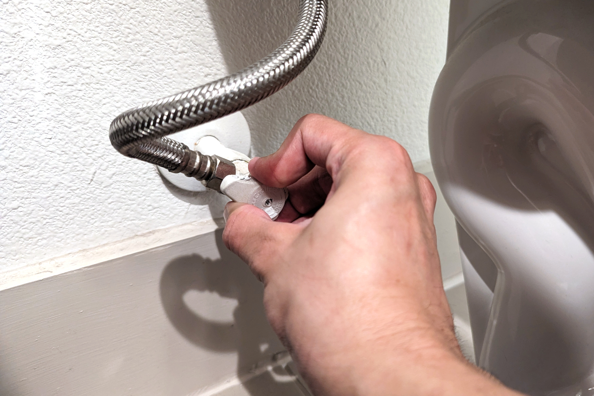 A person turning a toilet water supply valve from the "on" to the "off" position.