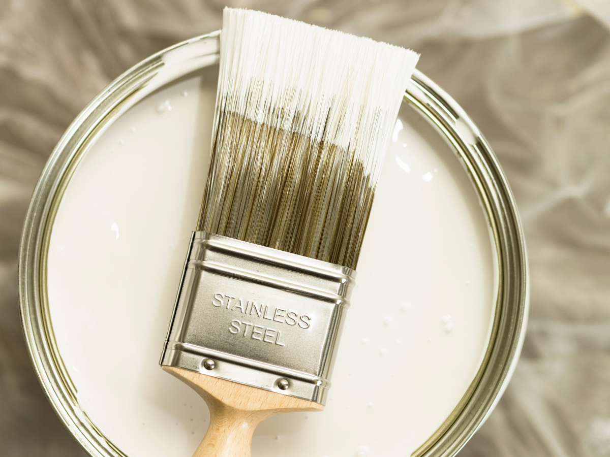 A loaded paint brush resting on an open can of white latex paint on a plastic drop cloth.