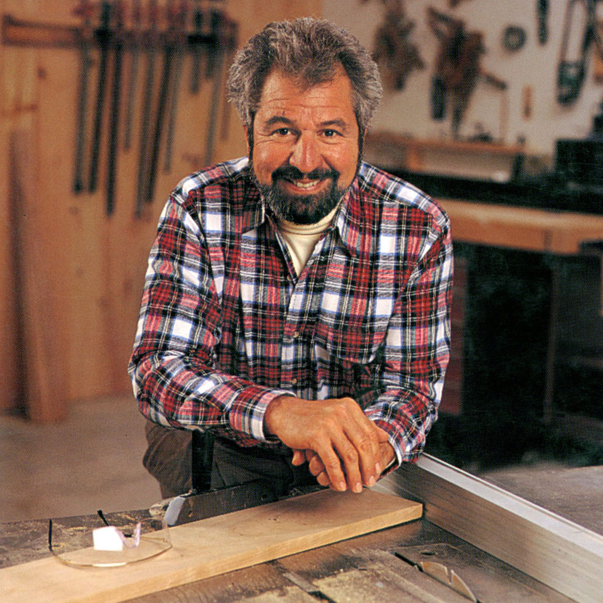 Bob Vila in a plaid shirt leans forward in a woodworking workshop.