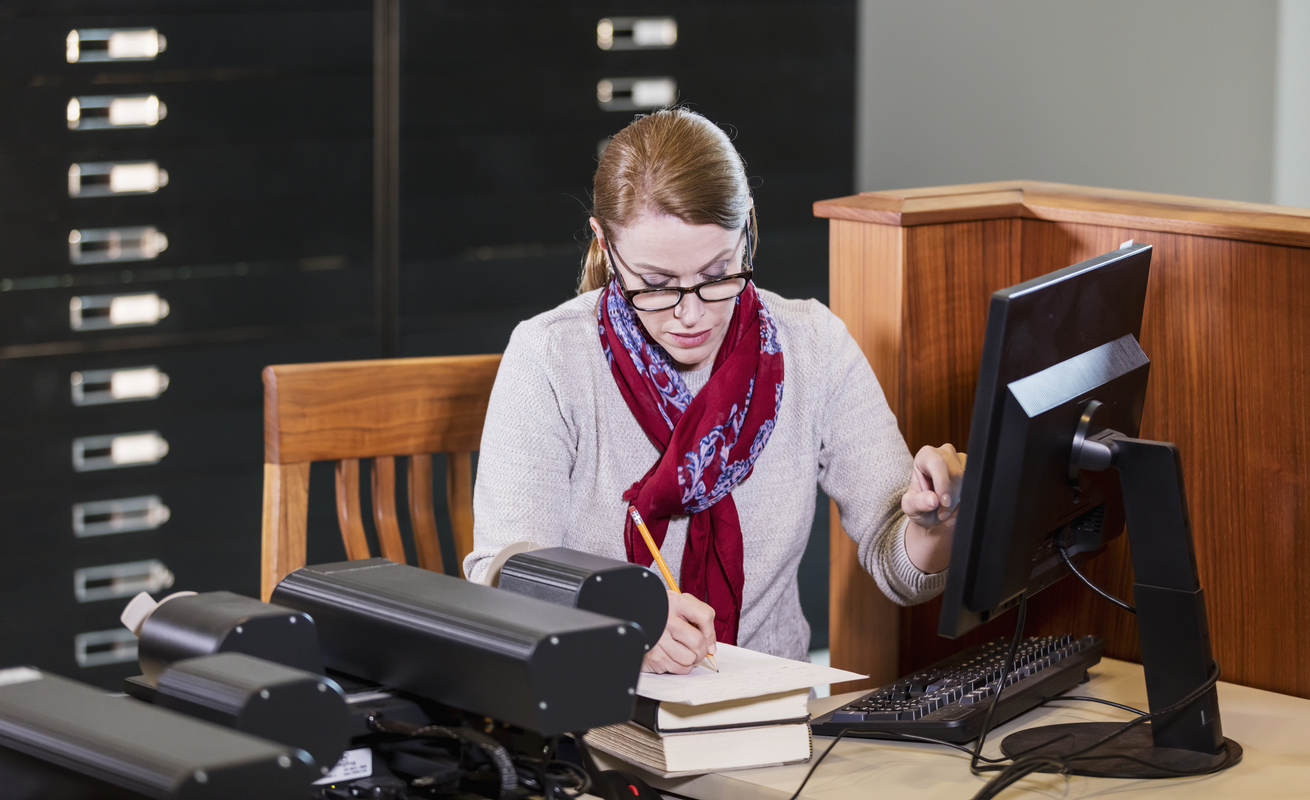 A mature woman in the library, using a computer to review historical documents stored on microfiche. She is writing notes.