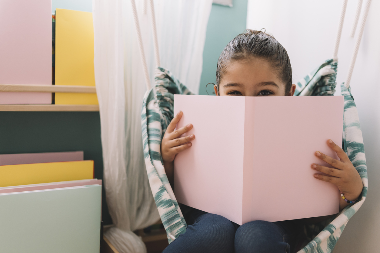 little girl hiding her face behind a pink book in a small reading corner