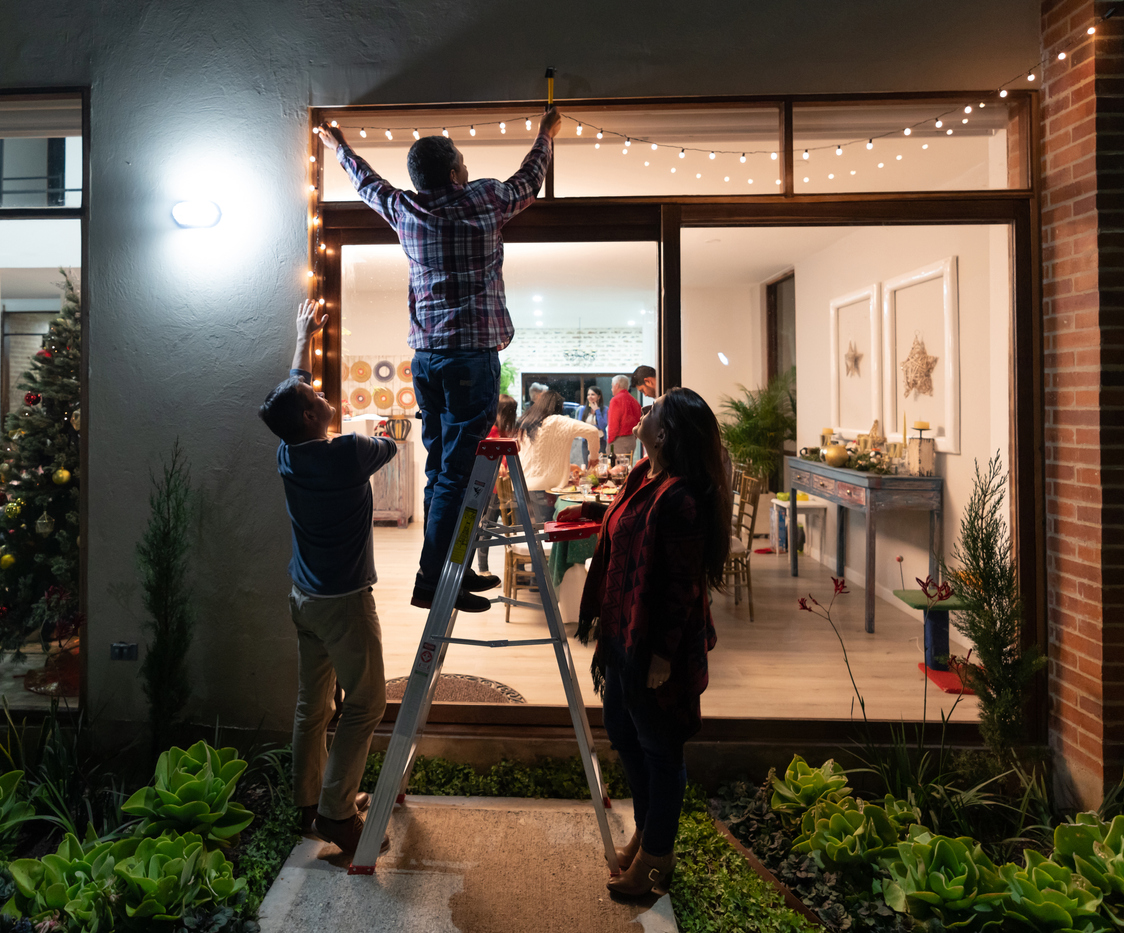 A man stands on a ladder hanging Christmas lights in the doorway with two family members watching and a view through sliding doors into the living room full of people.