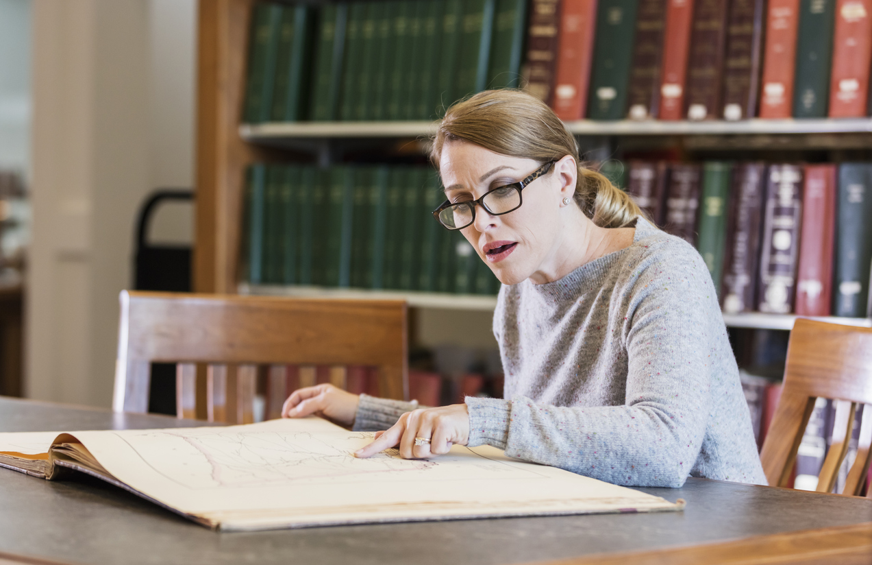 A mature woman in her 40s doing research in the library, sitting at a table looking at a large old book.