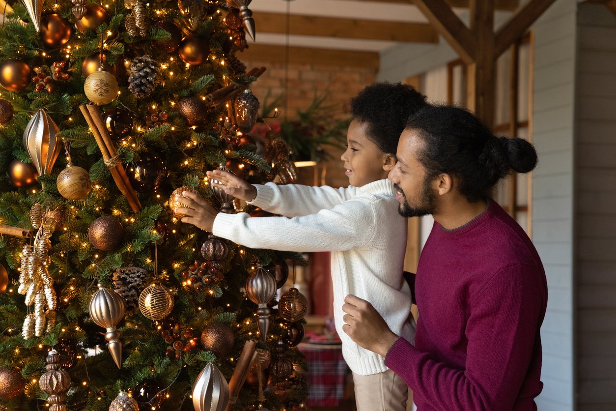 A father holds his son up to let him put an ornament on a Christmas tree in their living room.