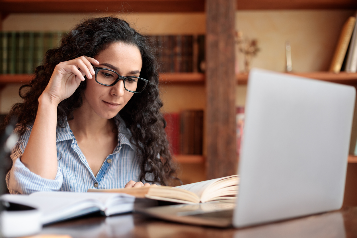 Young lady reading book sitting at desk touching glasses