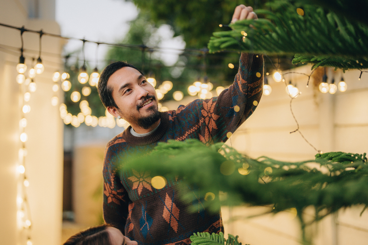 A man in a Christmas sweater hangs Christmas lights in his backyard.