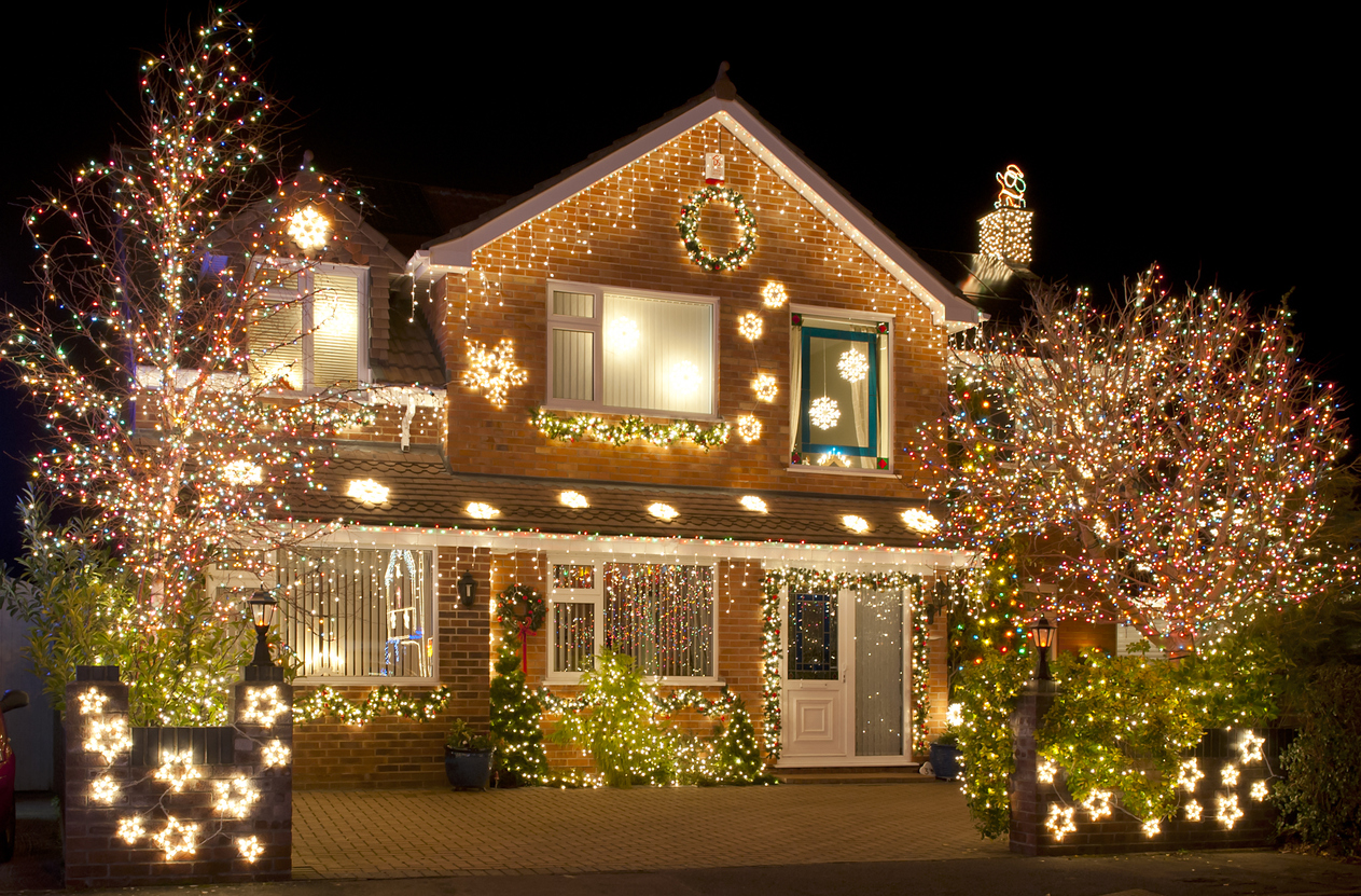 A stately brick house is decorated with bright white Christmas lights hanging around the porch and lining the roof and trees.