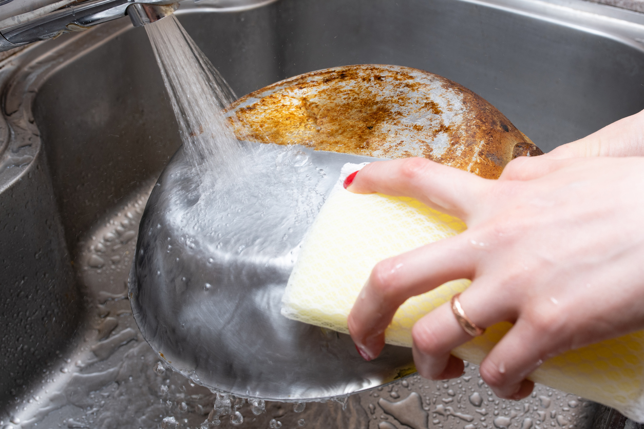 Woman with red nails and wedding band washes aluminum pan in the sink with yellow sponge and soap.