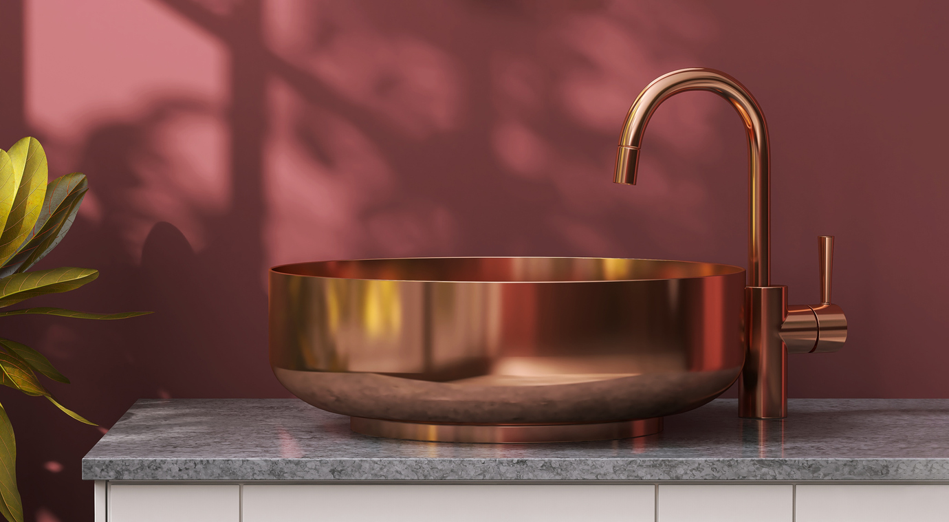 A copper sink and faucet is mounted on a granite counter with a maroon wall background.