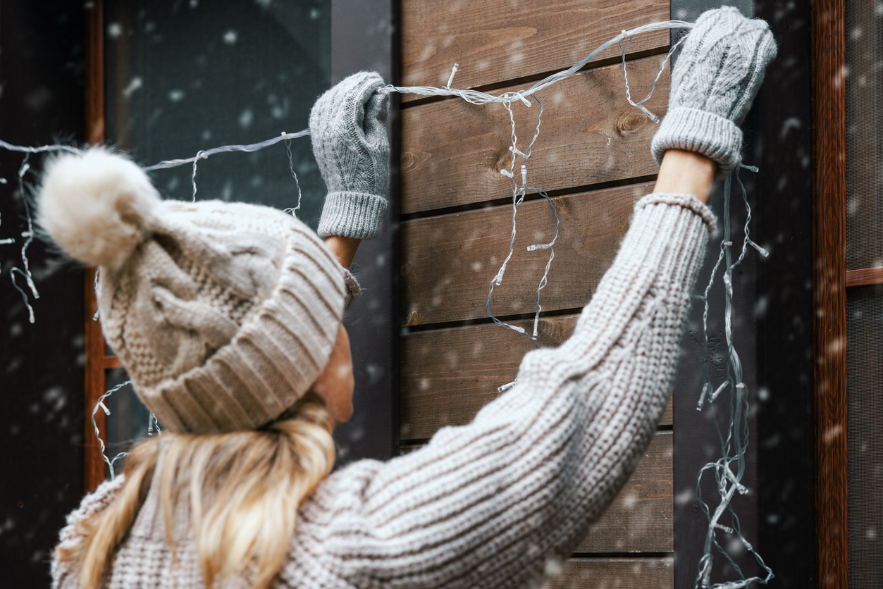A woman in a grey winter hat holds string lights up to the exterior wall and windows to hang them.