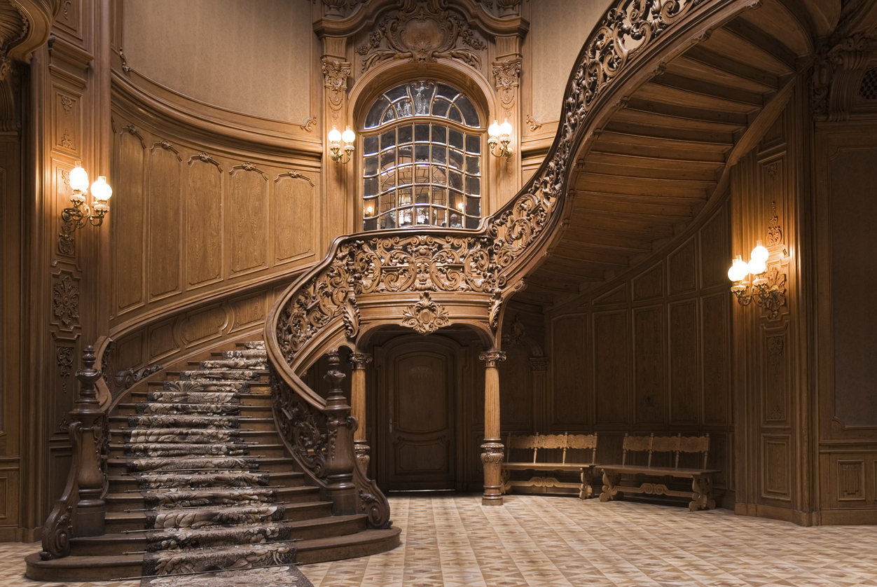 An-elaborately-carved-millwork-staircase-winds-around-in-a-wood-paneled-entry-hall.