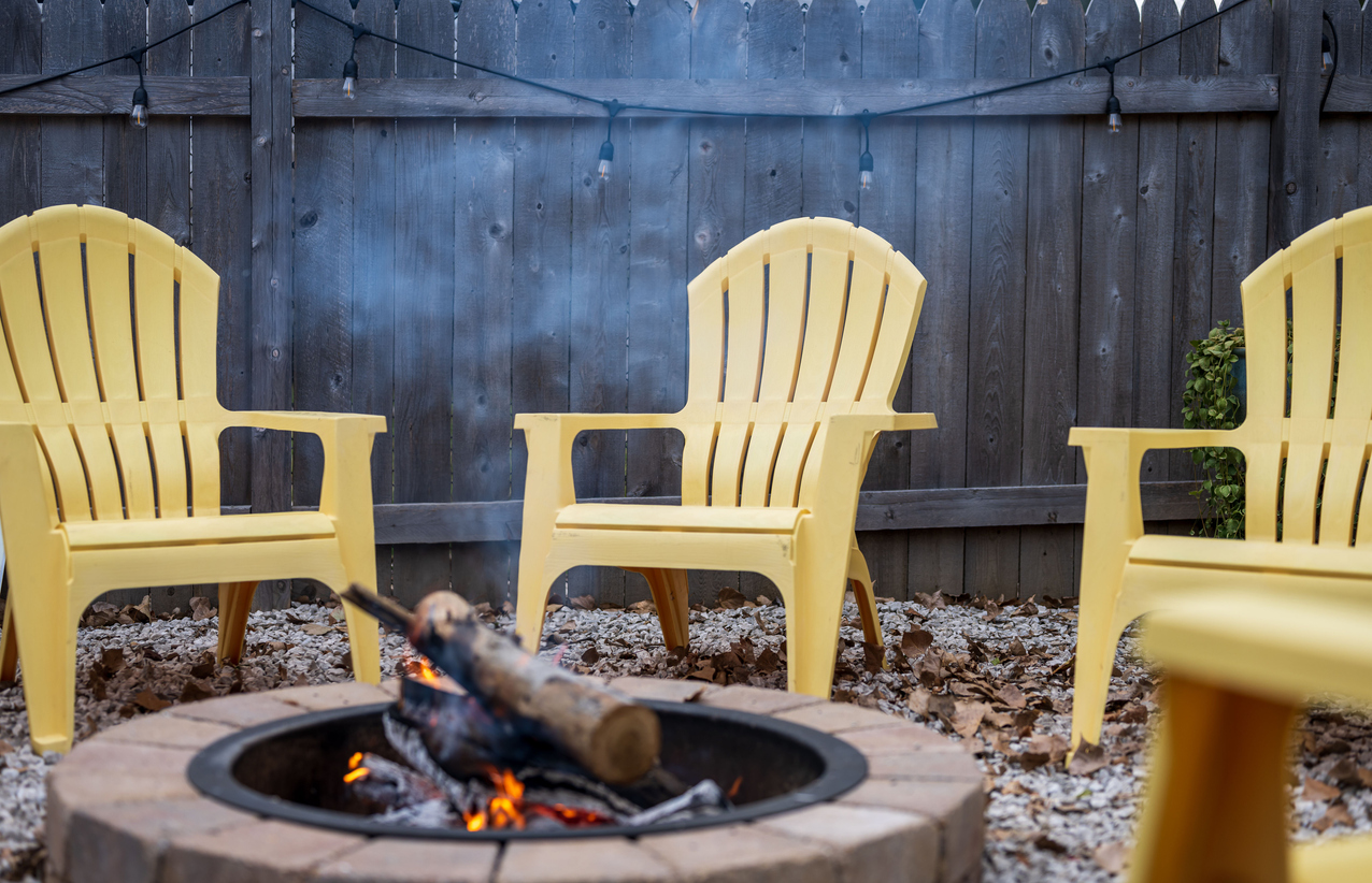 A fire pit with fire-burning logs, surrounded by yellow plastic chairs, in the backyard covered with fallen foliage, and fenced with a wooden fence