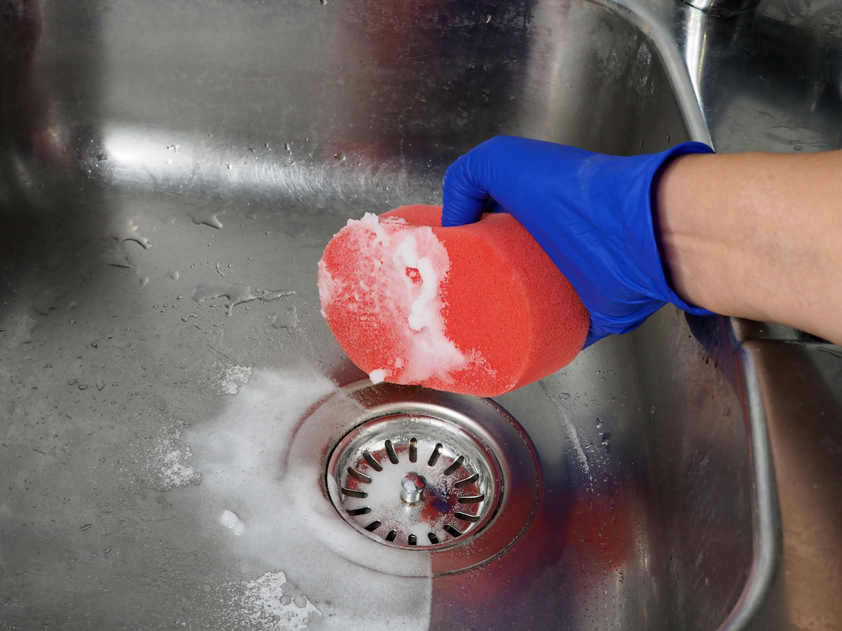 Person with blue gloves uses red sponge, baking soda, and vinegar to scrub stainless steel sink.