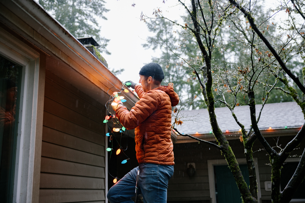 A man in an orange coat hangs Christmas lights on his house on a cloudy day.