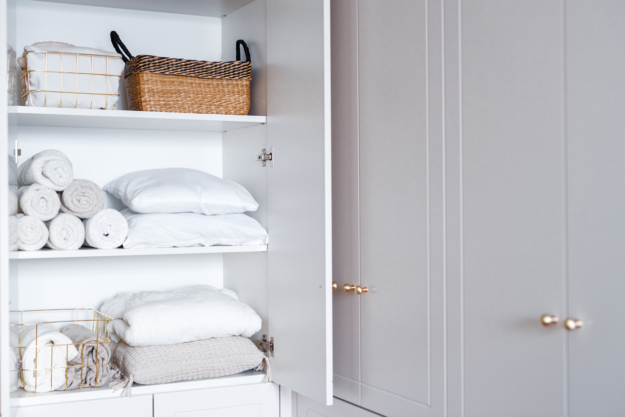 White laundry, pillows, towels organized and folded in baskets on shelves in white opened linen closet in the bathroom.