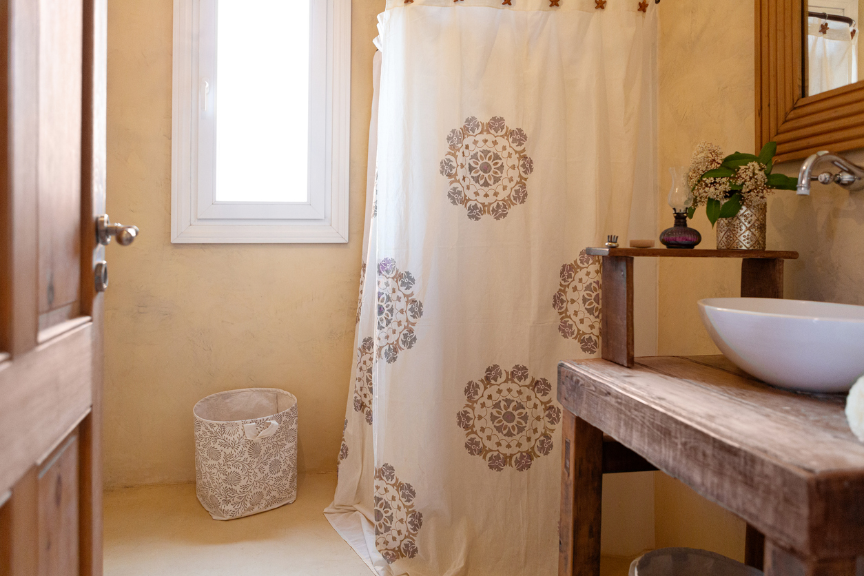 A wooden door is open to show a beige and brown bathroom with wood vanity and a shower curtain around the standing shower.