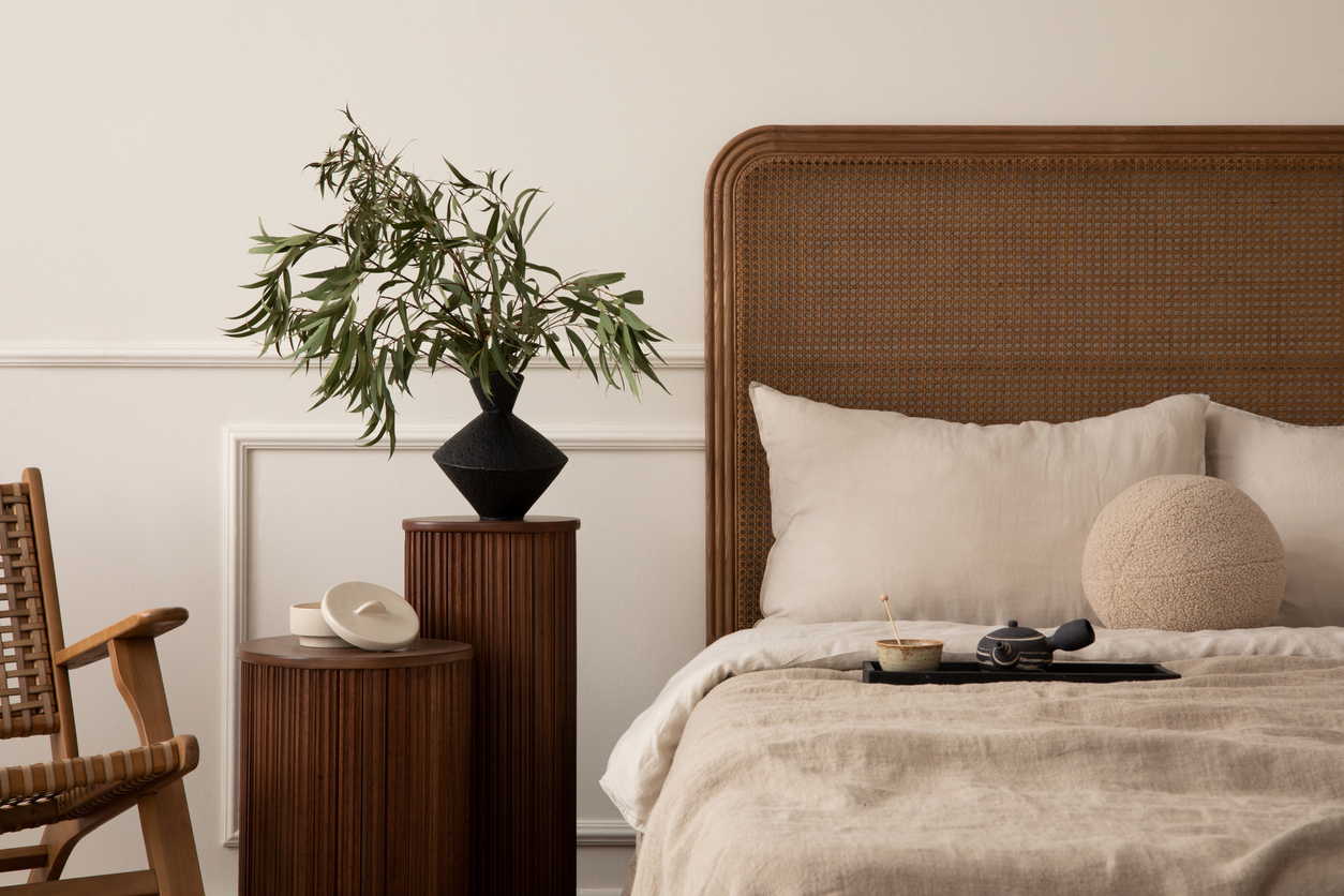 A bedroom with a wooden bed frame and rocking chair, linen duvet and a plant at the bedside table.