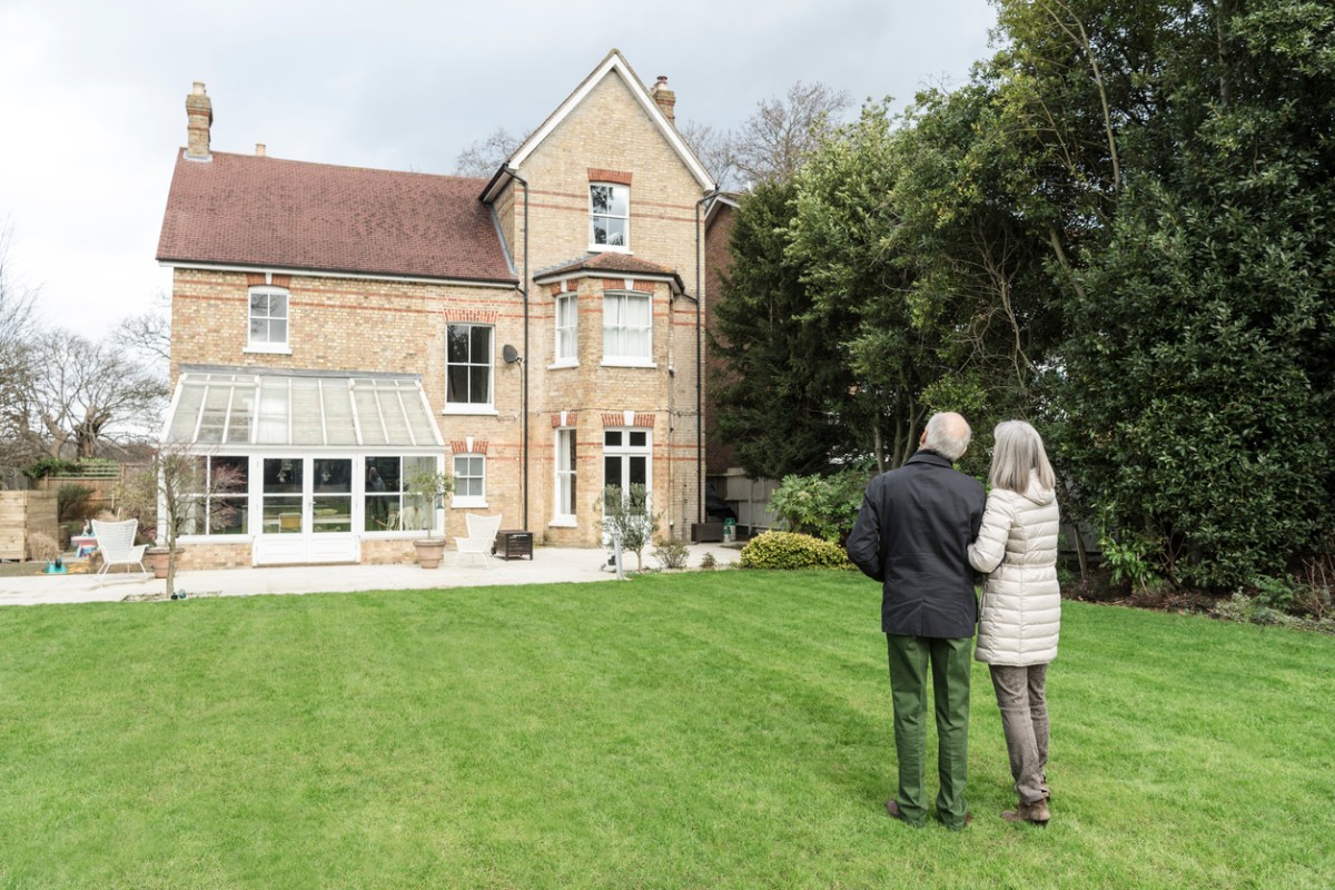 Two senior adults looking at the back of their house standing in the back garden on the grass. Senior man and woman arm in arm looking at home.