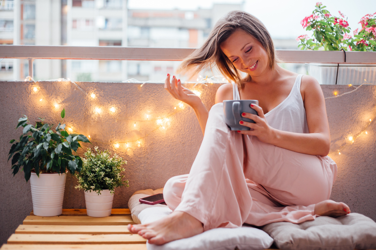 A-woman-enjoys-her-coffee-on-a-balcony-with-string-lights-and-potted-flowers.