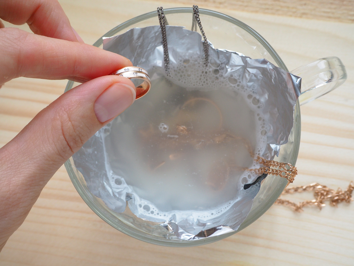 A woman's hand places a ring into a small glass teacup containing water with other pieces of jewelry being cleaned.