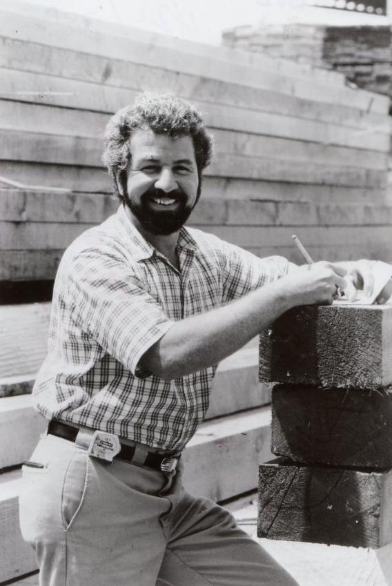 Bob Vila in a plaid shirt and full hair and beard write something down on the construction site.
