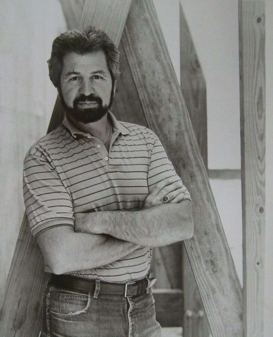 Bob Vila dressed in a polo and with his trademark beard leans against a home in construction.