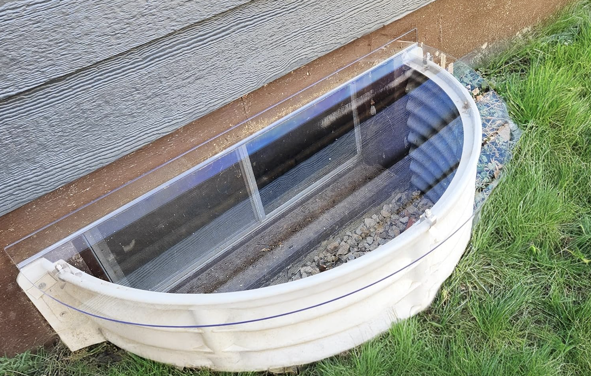 A deep basement window well with a clear window well cover protecting the area from rain. 