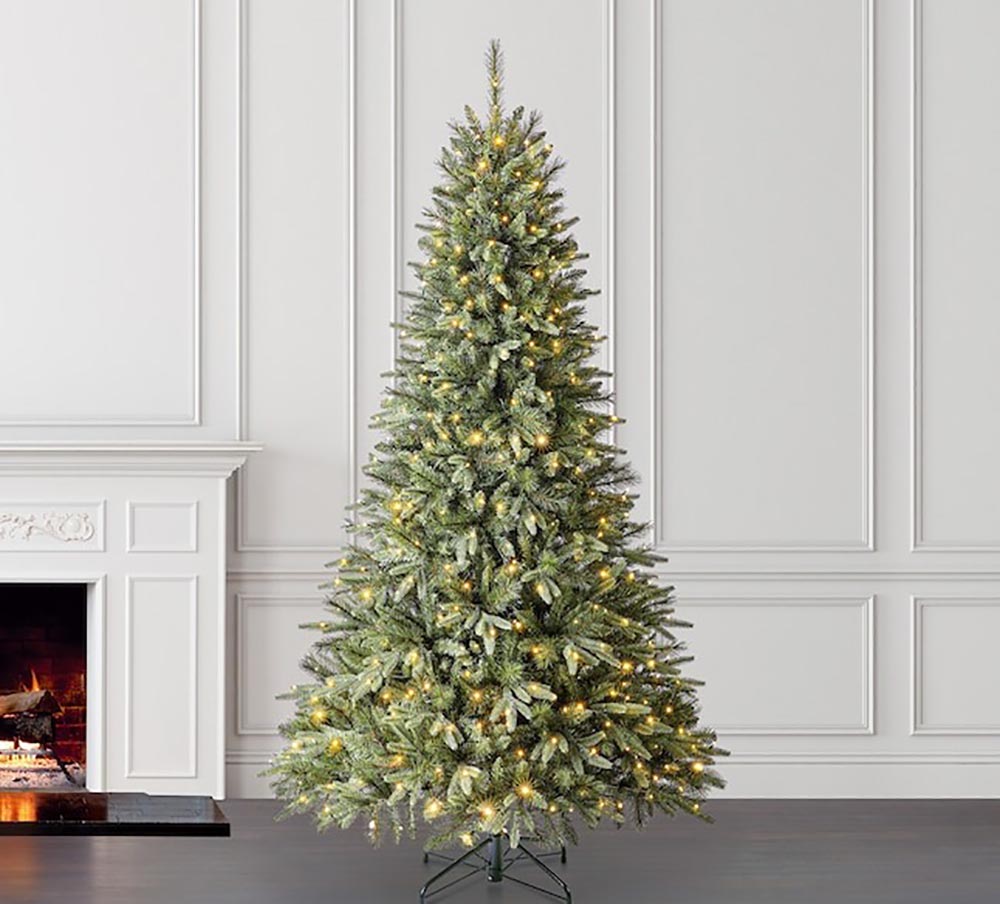 The Holiday Living Brighton Spruce Prelit Christmas Tree lit with white lights next to a white wall and white fireplace with a fire burning.