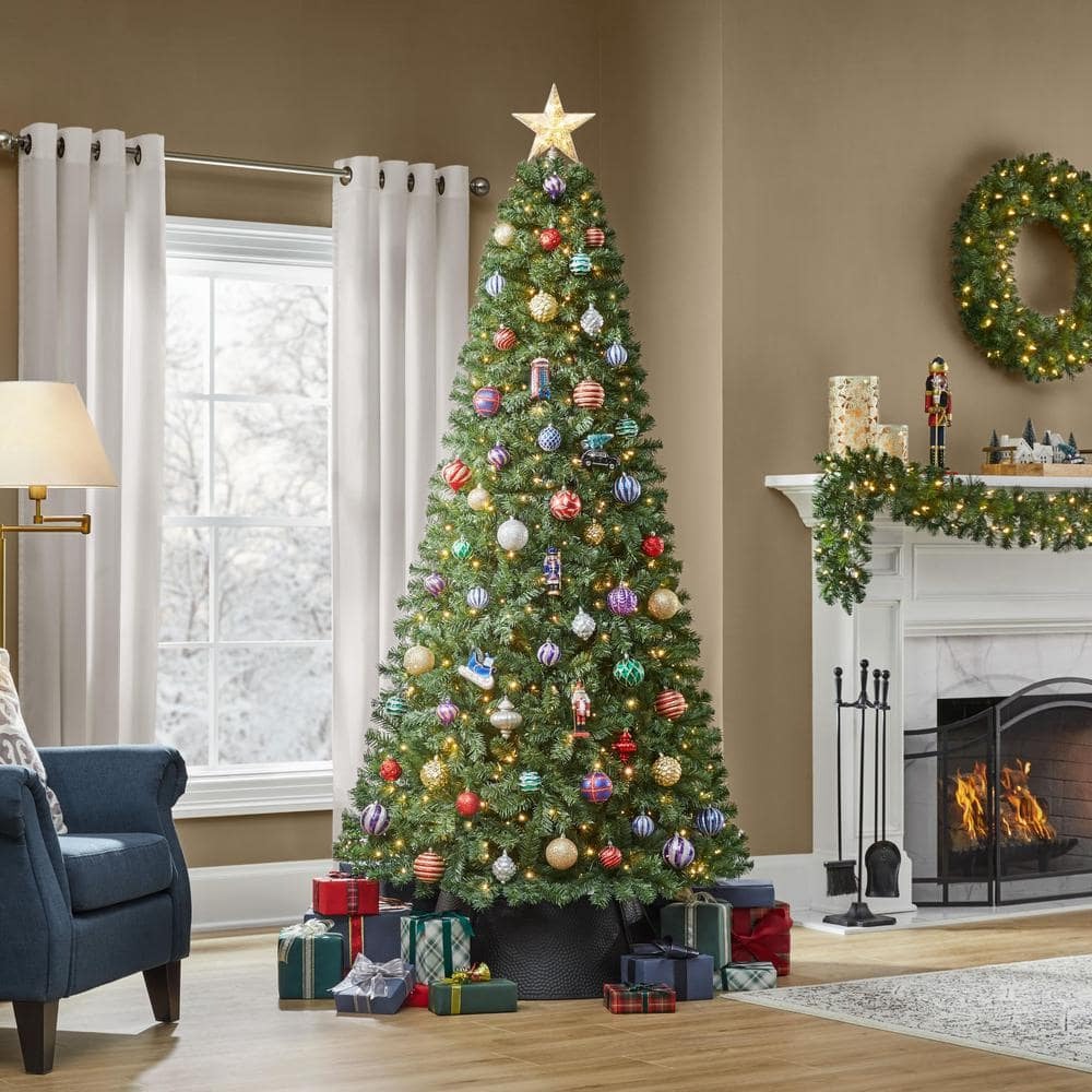 The Home Accents Holiday Prelit LED Festive Pine Tree decorated with ornaments and a star tree topper with presents underneath and a decorated fireplace in the background.