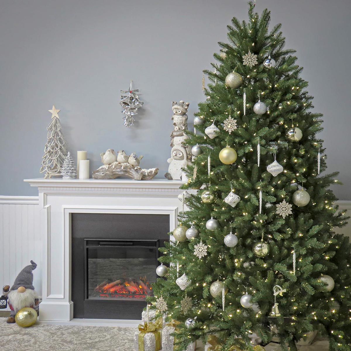 The National Tree Company Dunhill Fir Christmas Tree decorated with silver and gold ornaments and set up in front of a fire burning in a fireplace.