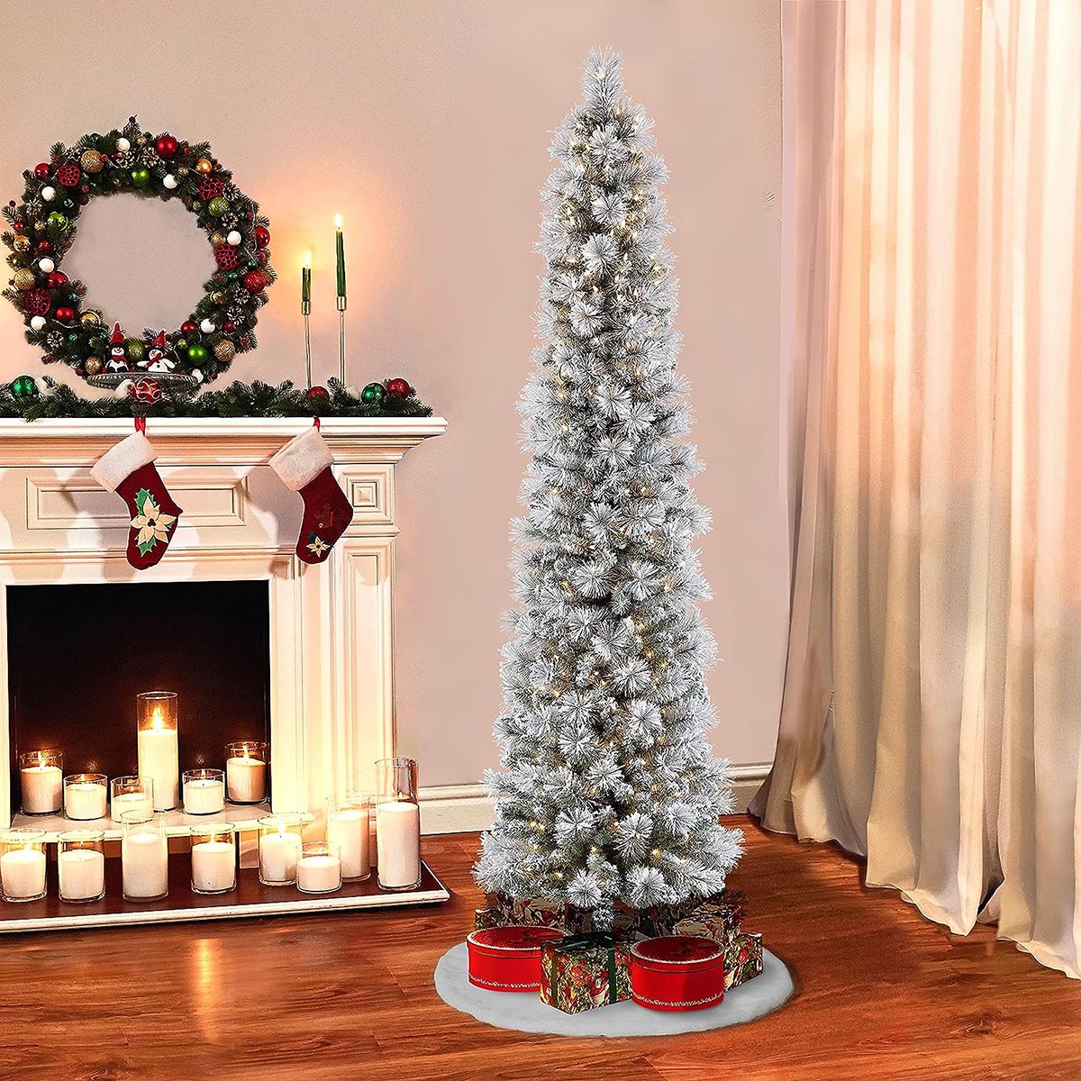 The Puleo International Prelit Flocked Pencil Tree with presents underneath while set up next to a fireplace filled with lit candles and decorated with a wreath and stockings.