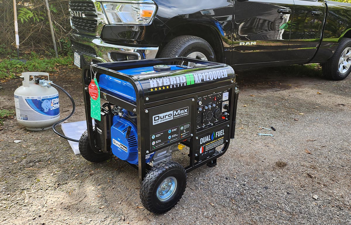 The DuroMax XP12000EH Generator on a gravel pad and hooked up to a propane tank with a truck in the background.